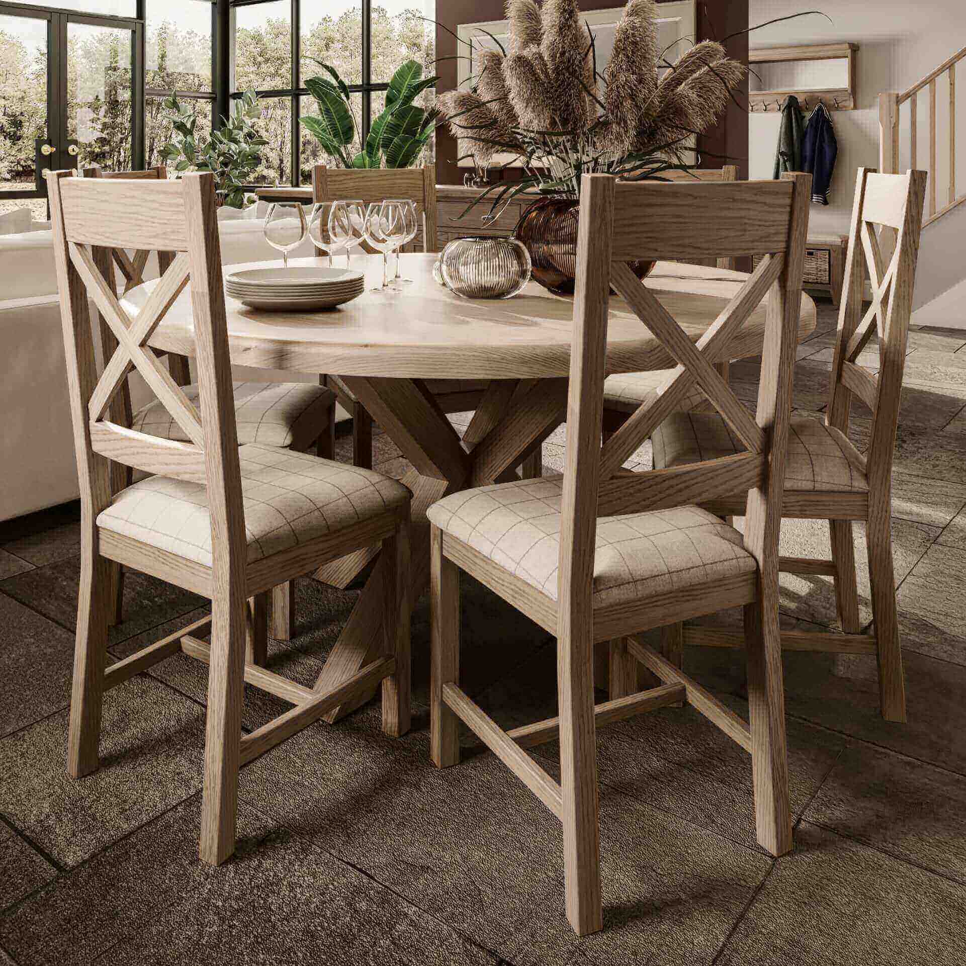 Large oak round dining table set with 6 cross back dining chairs around shown inside a cottage interior. Table is decorated with vase, candle and cutlery