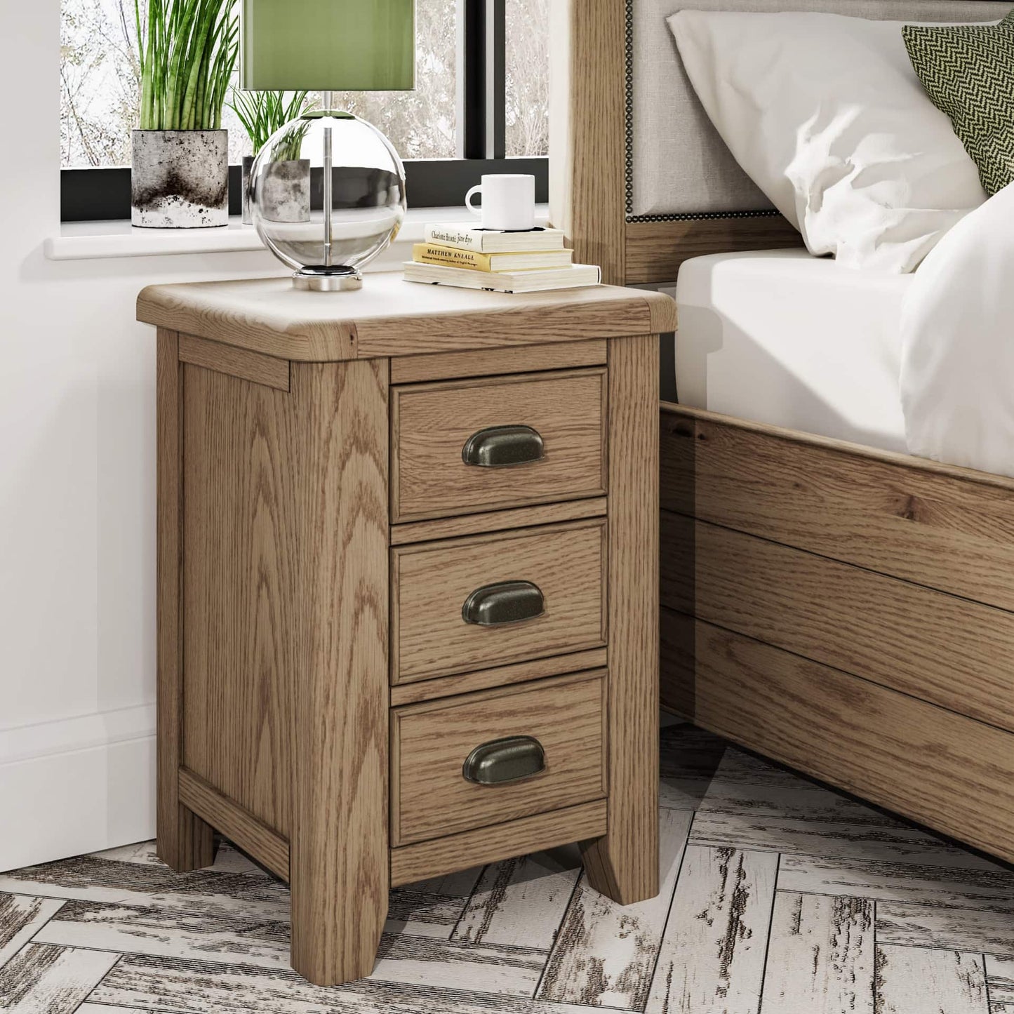 Oak bedside table with 3 drawers in medium size is shown in a room setting next to a bed