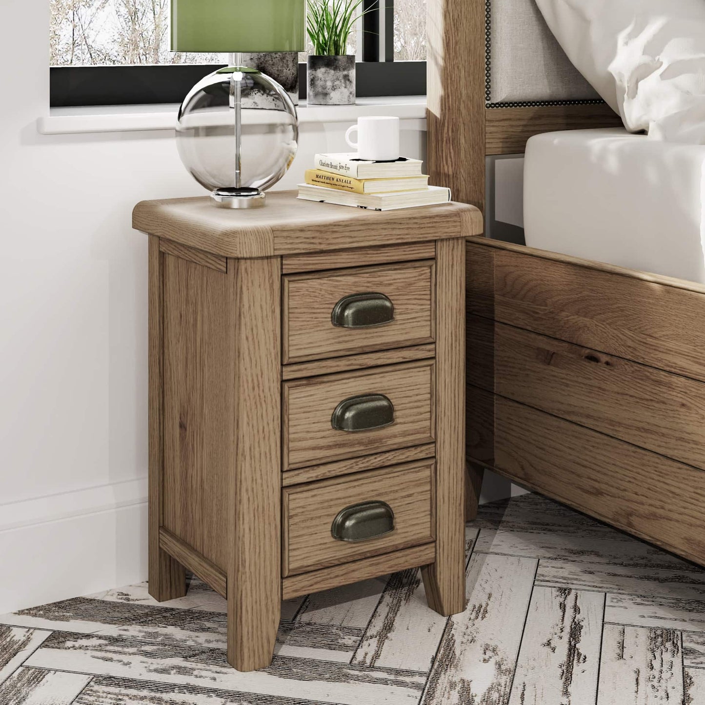 Oak bedside table with 3 drawers in small size is shown in a room setting next to a bed