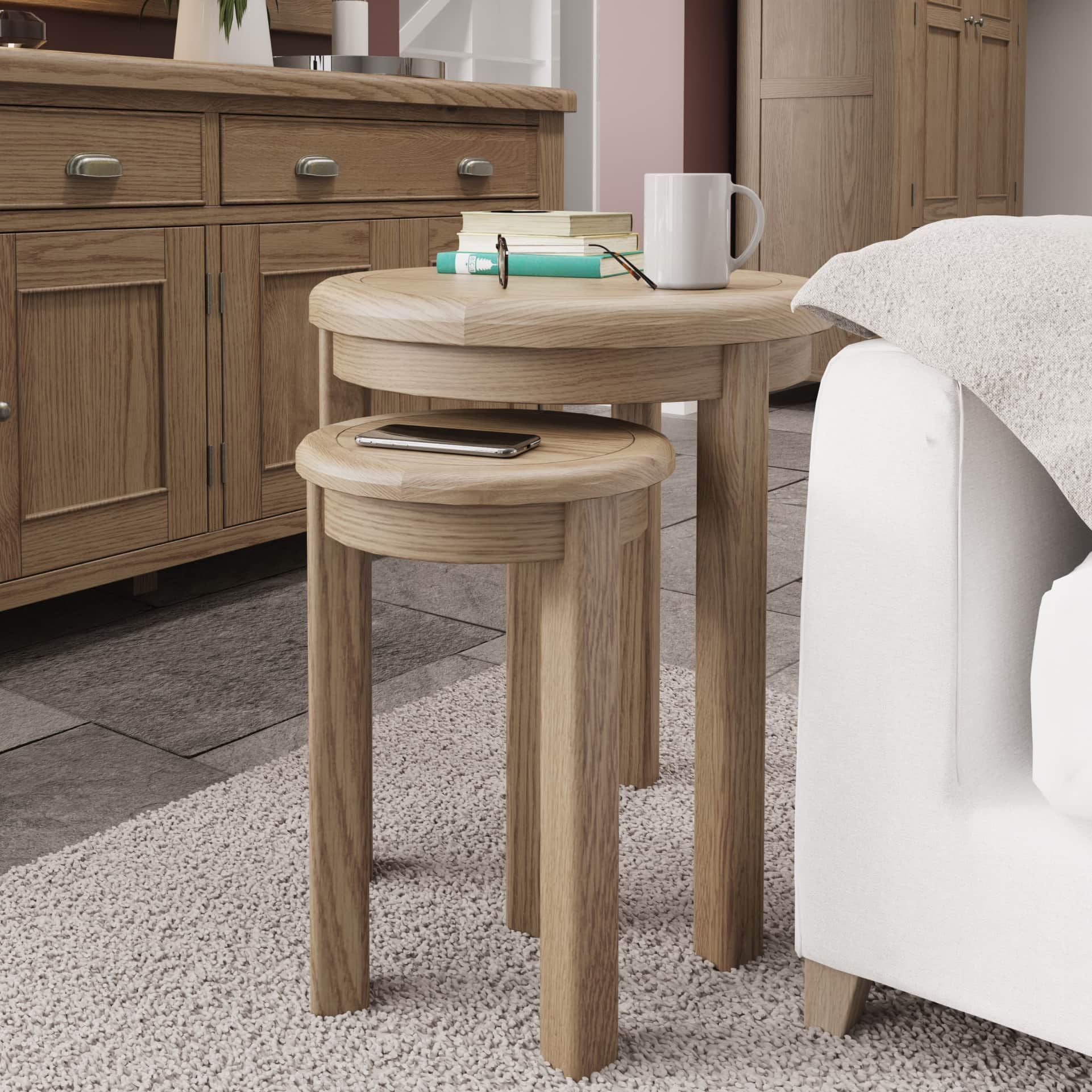 Round oak nesting side tables are shown in a room setting positioned next to a sofa. A pair of nesting side tables is used to hold a mug, books, glasses and a phone