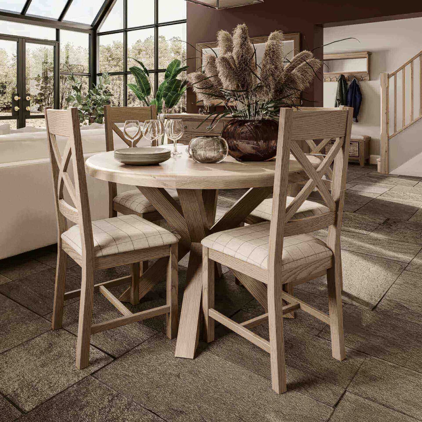 Small oak round dining table set with 4 cross back dining chairs around shown inside a cottage interior. Table is decorated with vase, candle and cutlery