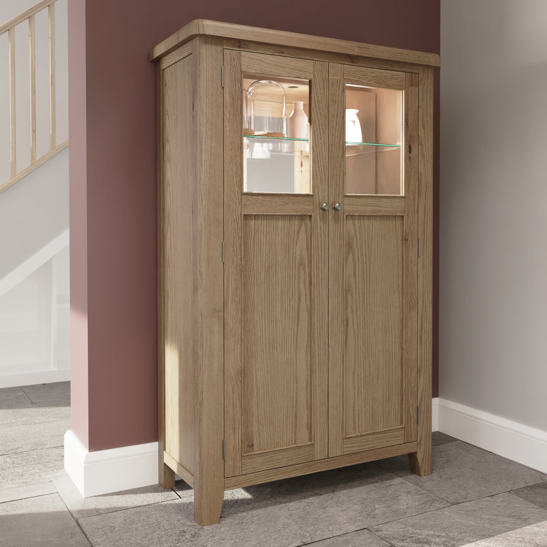 Oak tall wooden drinks cabinet is shown in room setting. The 2 upper display shelves behind glass exhibiting decor and vases with LED lights and mirrored back