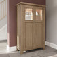 Oak tall wooden drinks cabinet is shown in room setting. The 2 upper display shelves behind glass exhibiting decor and vases with LED lights and mirrored back