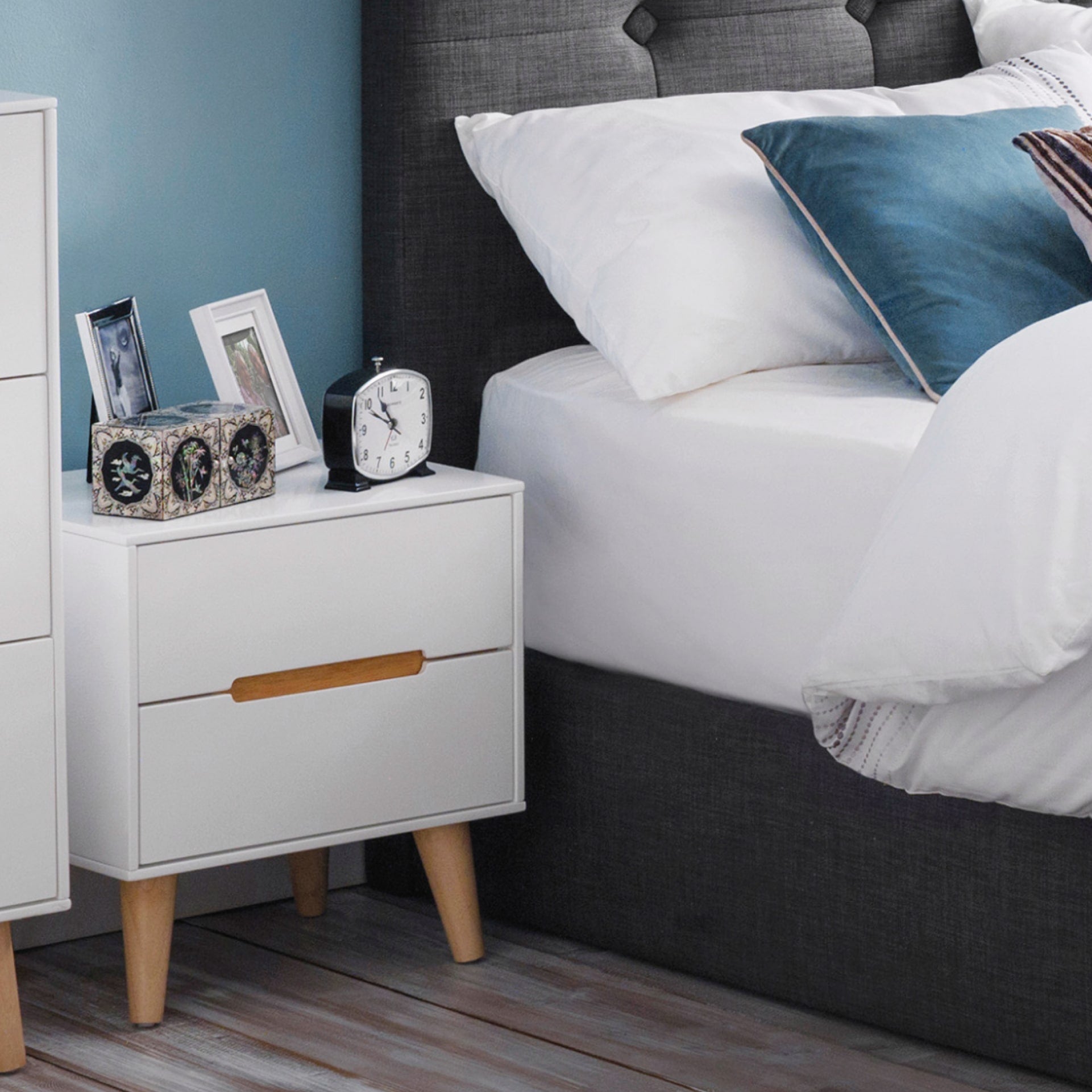 Alicia 2 drawer white bedside table is shown next to the bed with clock and accessories on top. Legs and recessed handles are in contrasting oak colour 