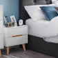 Alicia 2 drawer white bedside table is shown next to the bed with clock and accessories on top. Legs and recessed handles are in contrasting oak colour 