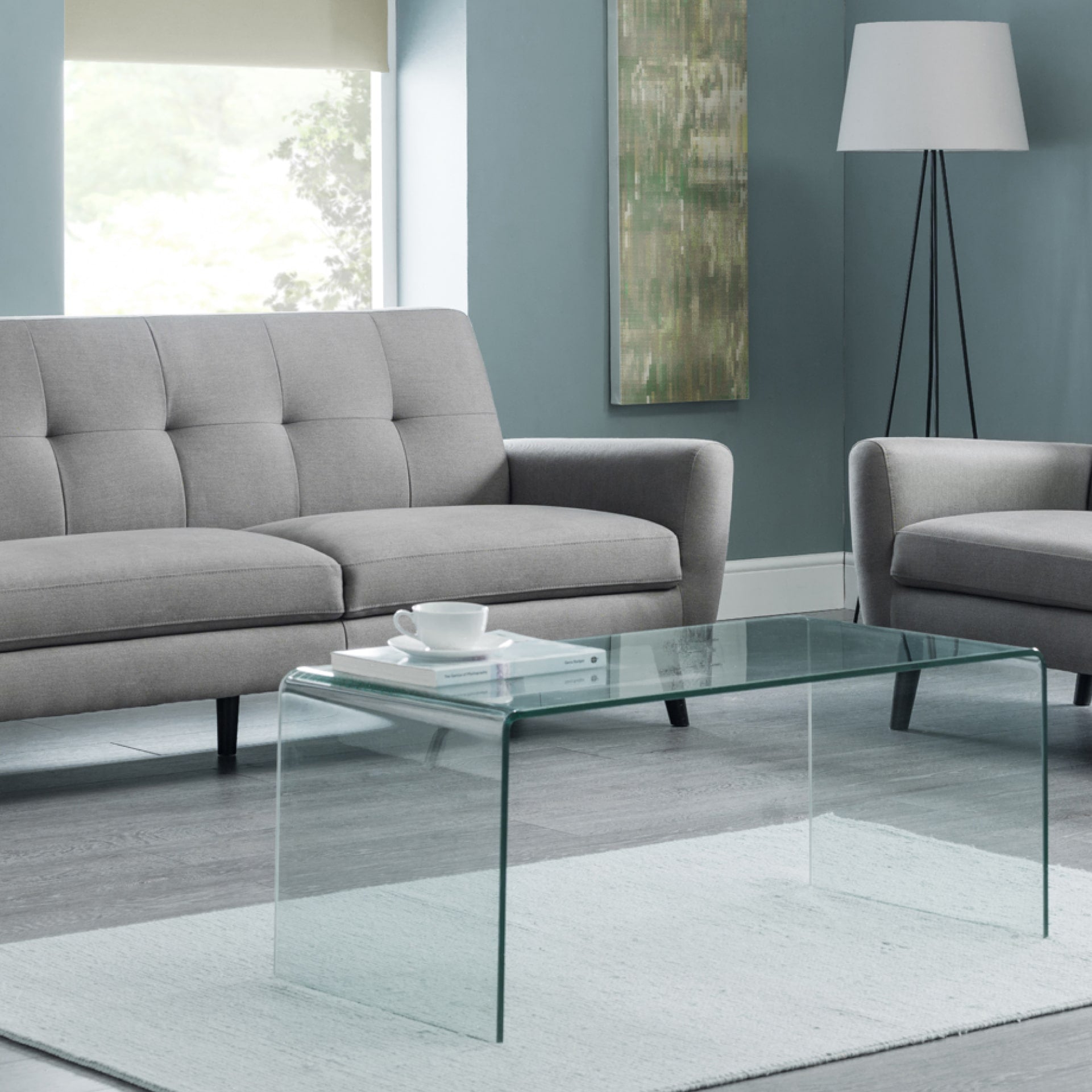Minimalistic glass coffee table is shown in living room setting standing on a carpet next to sofa and armchair set. 