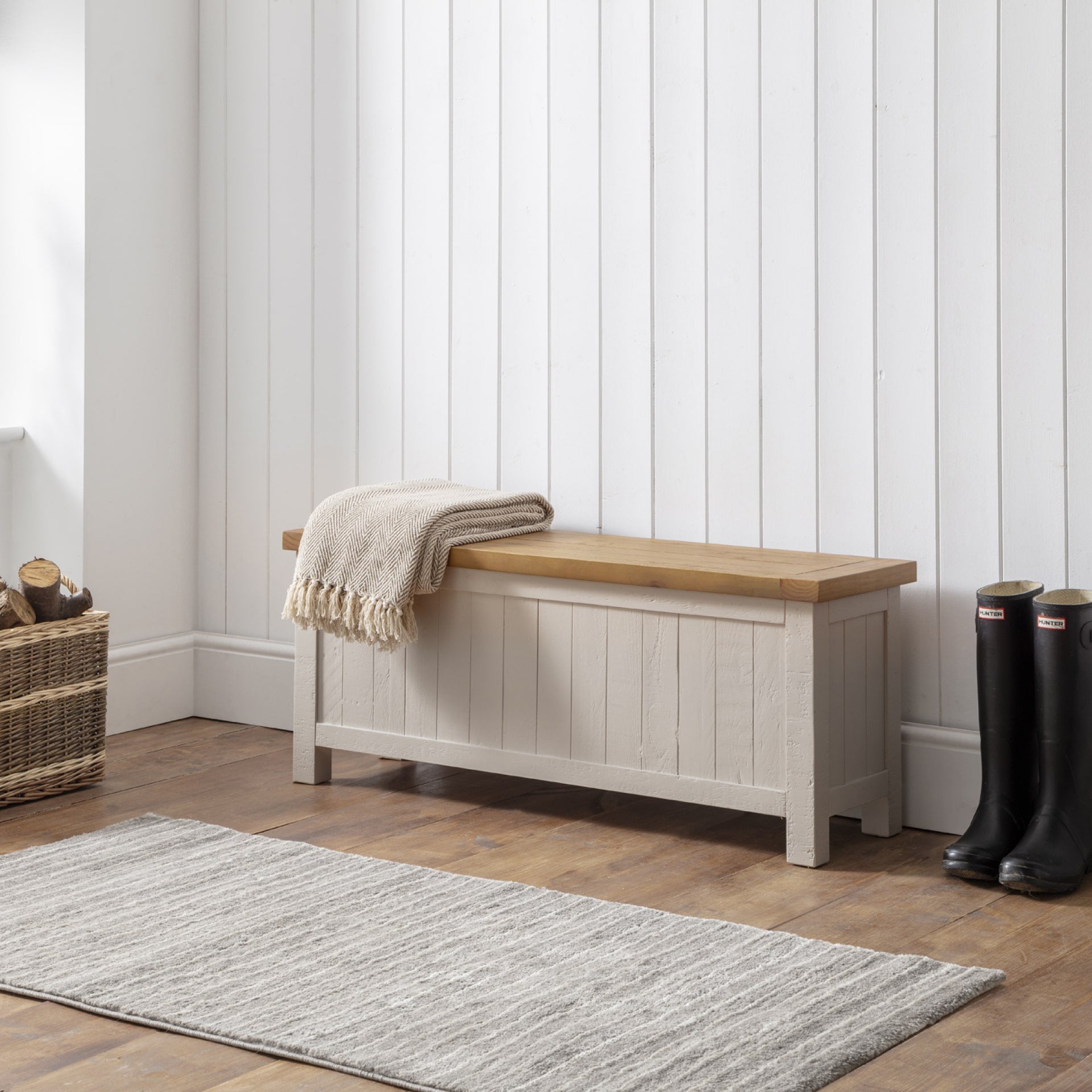 Grey storage bench is shown in an entryway / mudroom interior with fire wood, blanket and wellies around 
