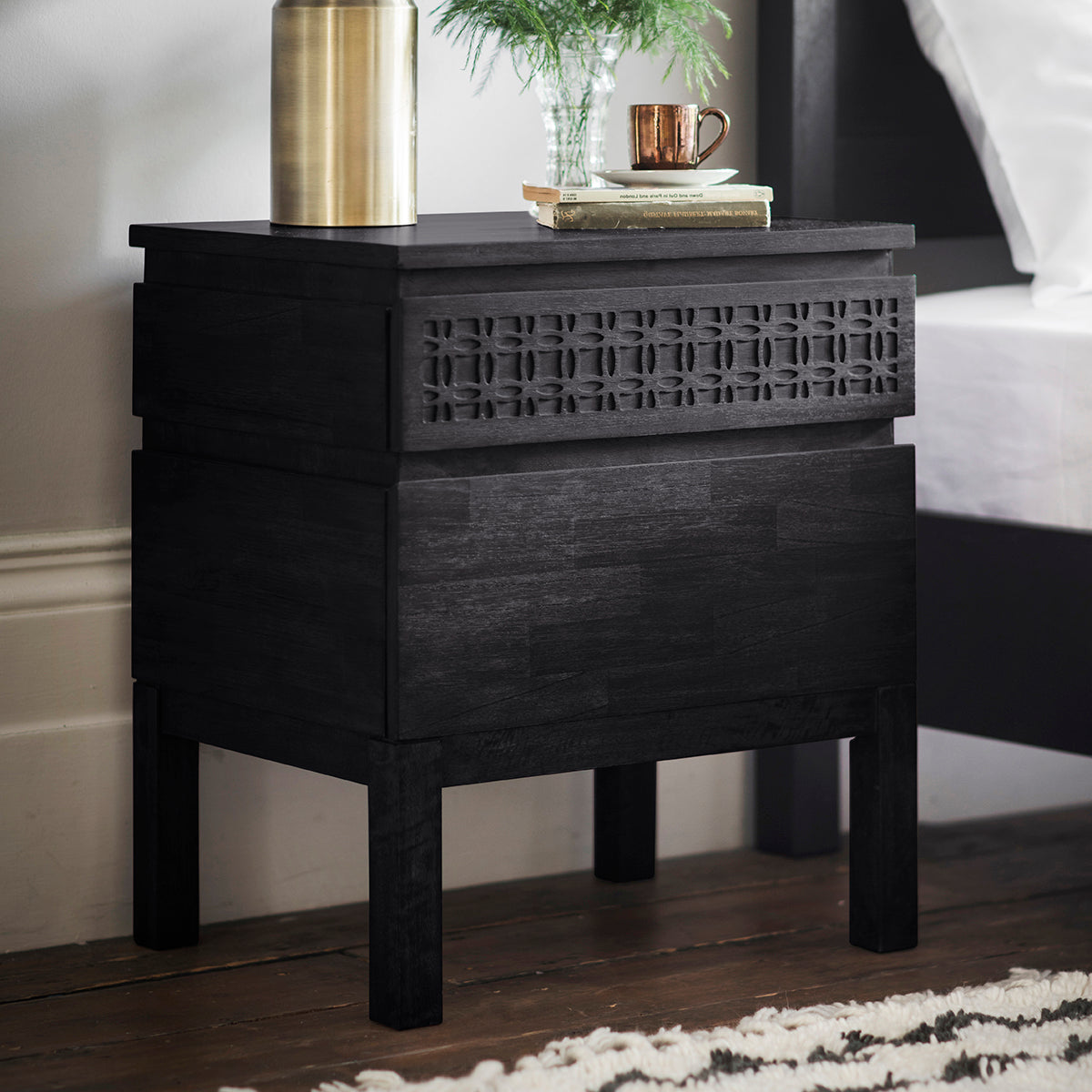 Modern nightstand with intricate detailing on the top drawer looks elegant and stunning in the bedroom setting with a vase, books and cup of coffee. The two drawers offer space for bedtime essentials. 