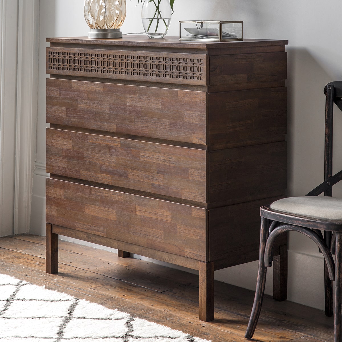 Magnificent brown chest with 4 drawers of storage is shown in the room setting with decor. The front of the top drawer has a stunning frieze of the blind fretwork and the drawers are made with mixed wood veneers put together in a mosaic manner shimmering in the light.